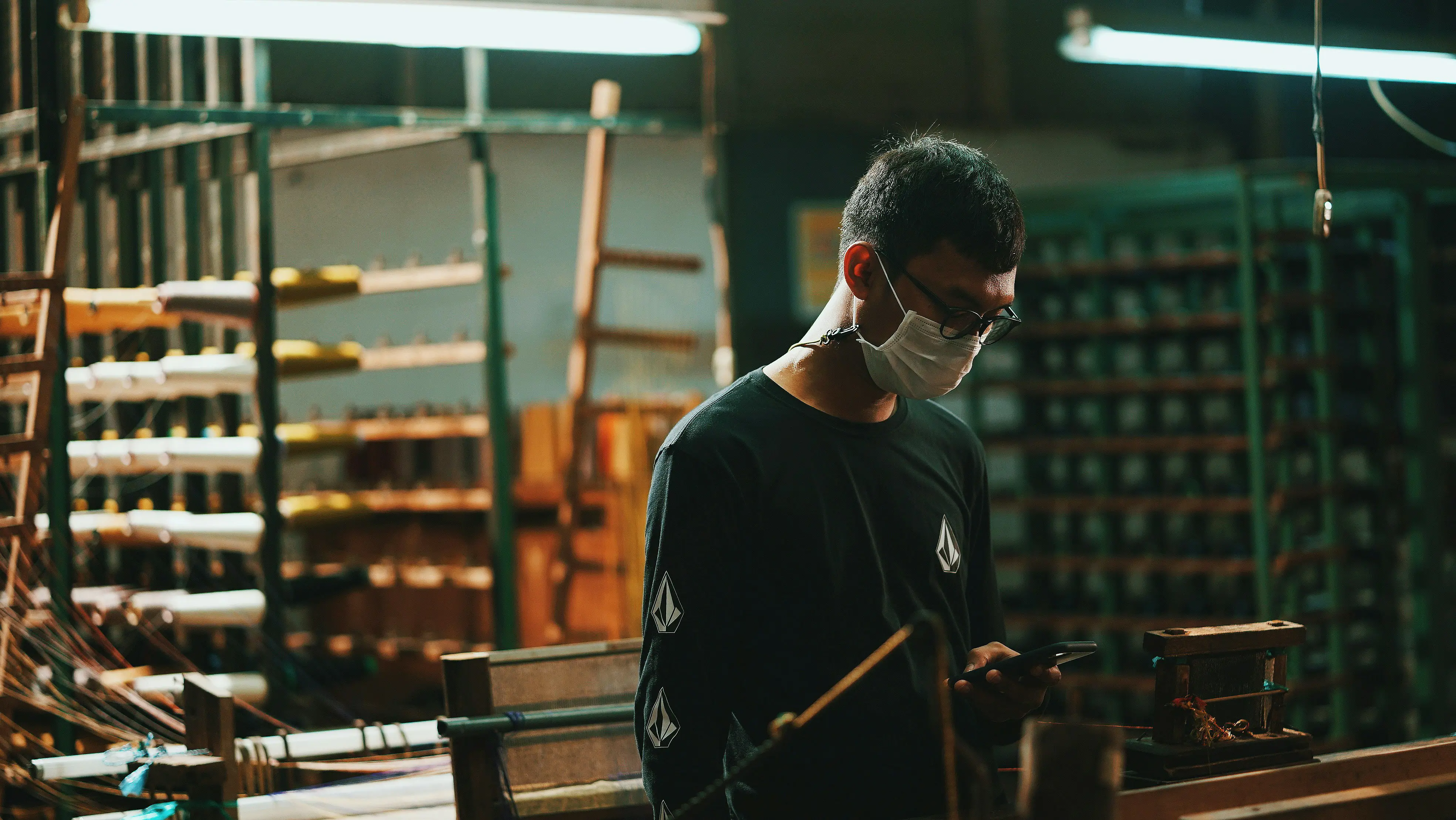 Man in manufacturing wearing a mask, looking at his phone near machinery.